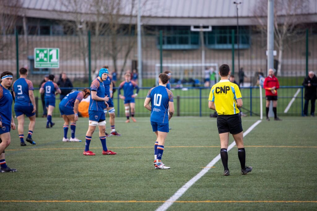 Action-packed rugby match with teams in blue and referee in a stadium, Manchester, UK.